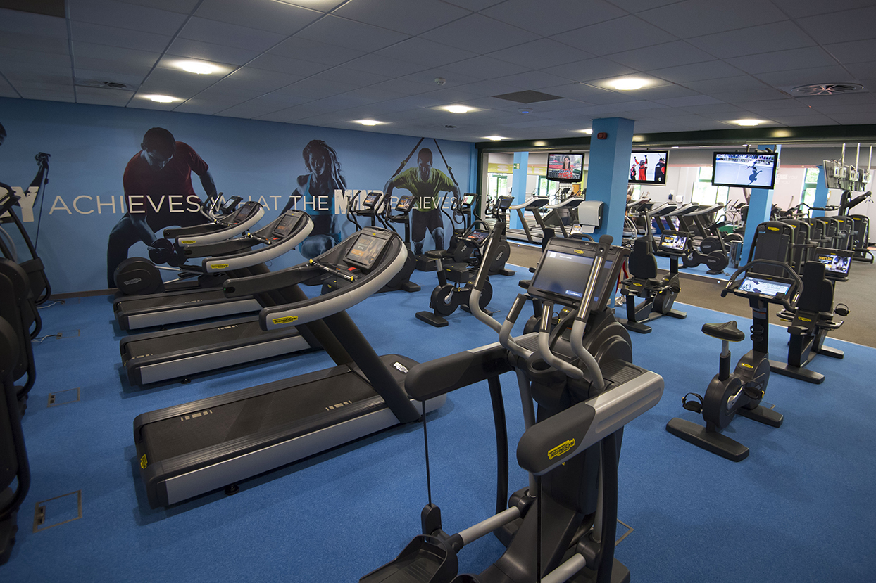 Gym interior with treadmills, stationary bikes, and exercise equipment on a blue floor, with motivational wall graphics.