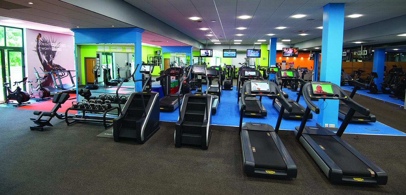 A spacious, brightly lit gym with treadmills, stationary bikes, dumbbells, and various fitness equipment.