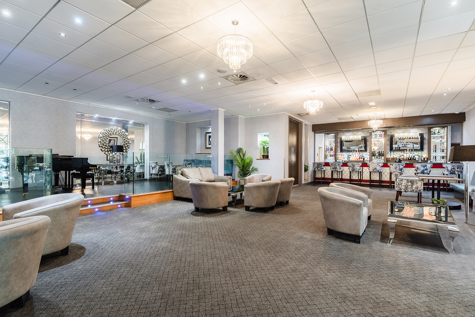 Spacious hotel lounge with armchairs, a stage, and a bar in the background, featuring chandeliers and modern decor.