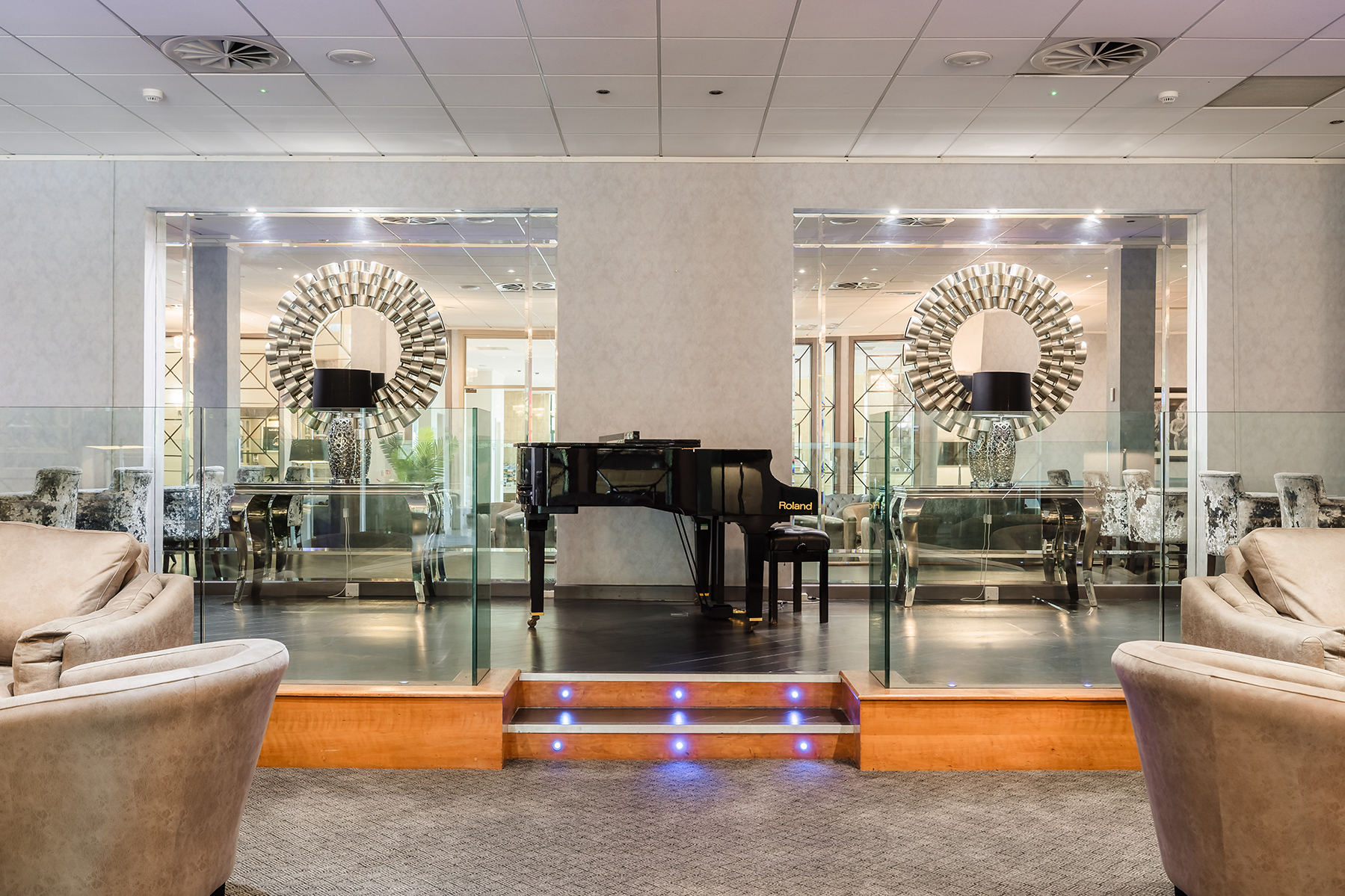 A grand piano is centered on a stage between two decorative mirrors in a modern lounge area with plush seating.
