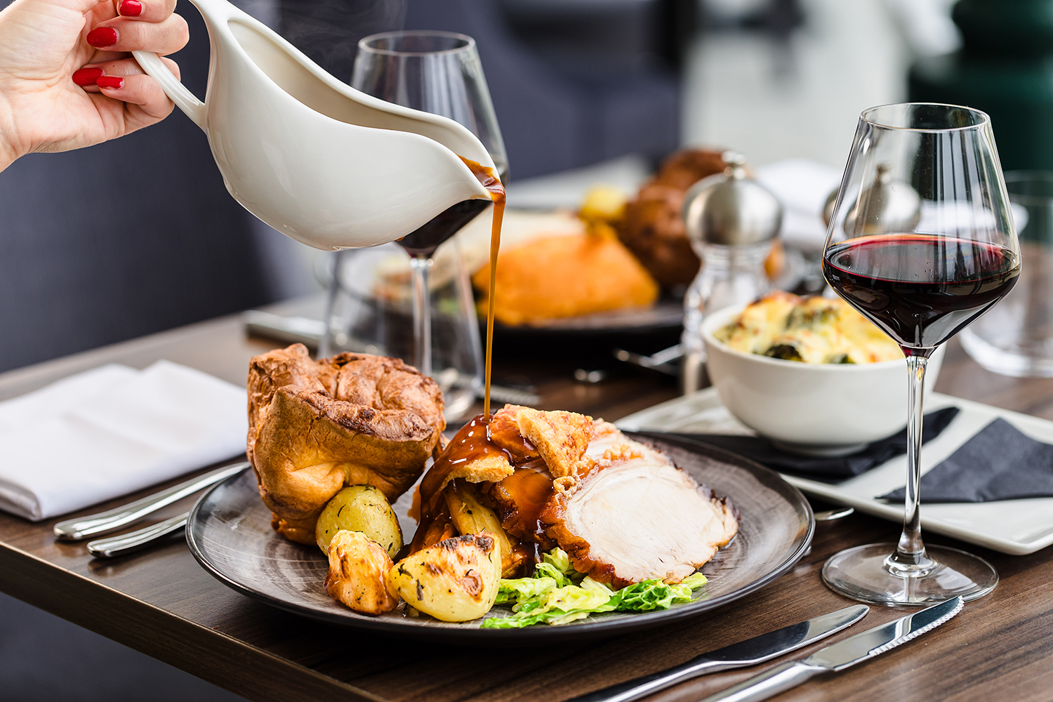 A hand pours gravy over a plated roast dinner with potatoes, vegetables, and a Yorkshire pudding, next to a glass of red wine.