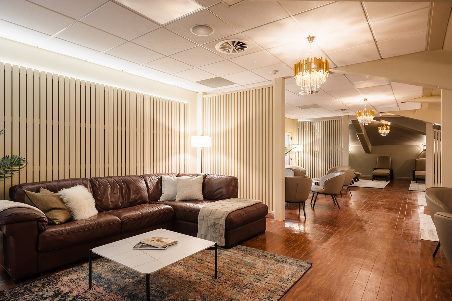 Spacious lounge with a brown leather sectional sofa, coffee table, armchairs, wooden floor, and ornate ceiling lights.