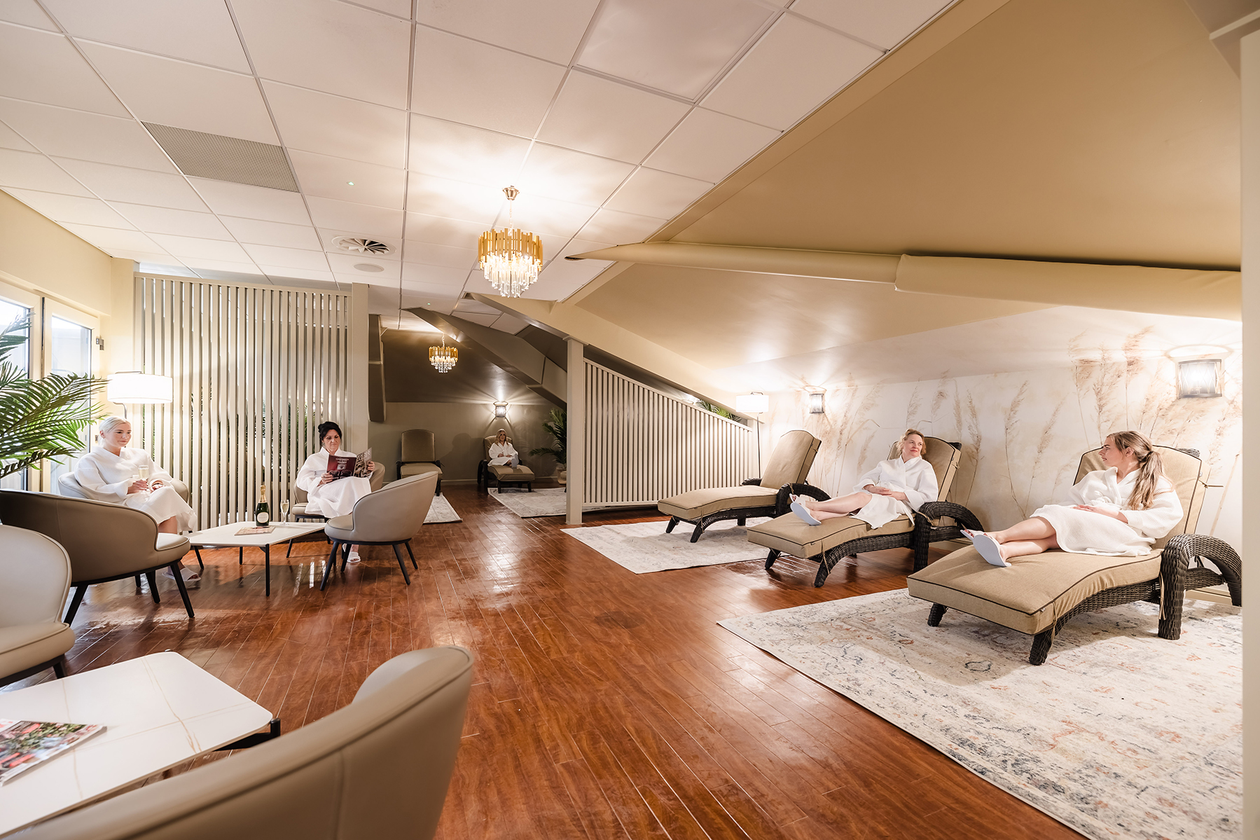 People in white robes relaxing on lounge chairs in a spa with warm lighting and wooden floors.