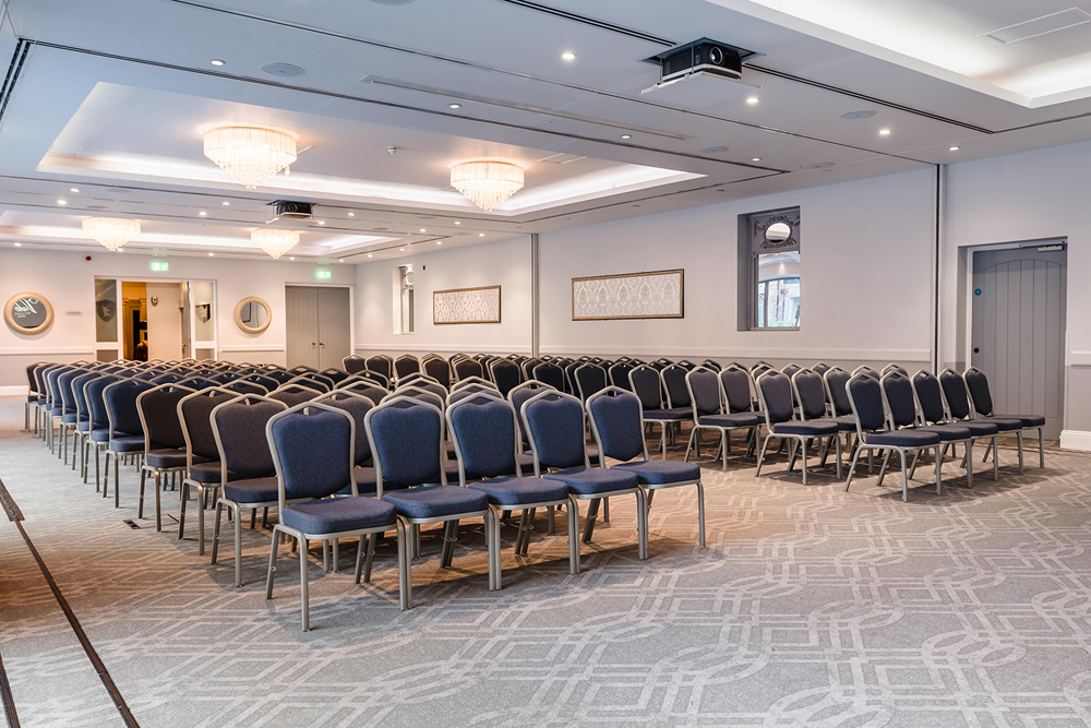 Empty conference room with rows of blue chairs, chandeliers, and patterned carpet.