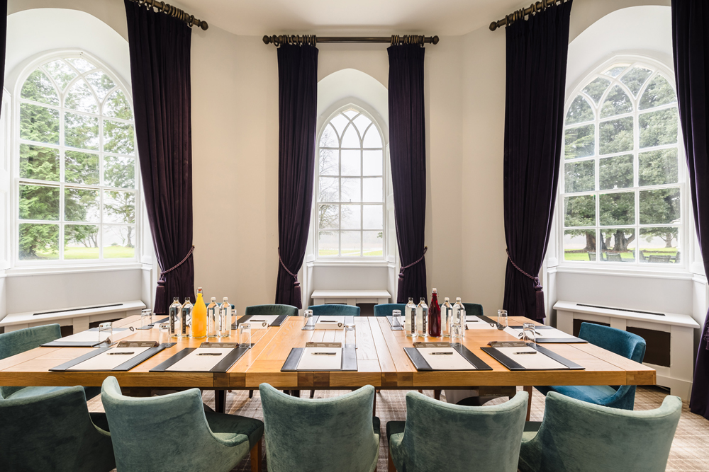 Conference room with a large wooden table set for a meeting, featuring chairs, notepads, pens, and drinks, surrounded by arched windows.