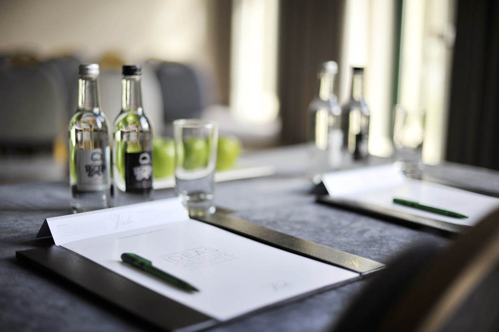 Close-up of a meeting table setup with notepads, pens, water bottles, glasses, and green apples on a gray surface.