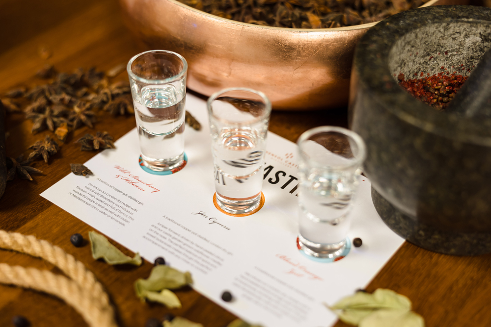 Three shot glasses on a tasting mat surrounded by spices, including star anise and bay leaves, with a mortar and pestle nearby.
