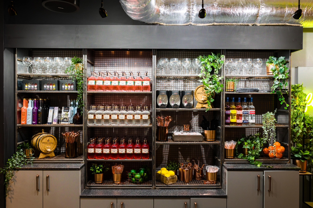 A bar shelf displays liquor bottles, glassware, bar tools, herbs, and citrus fruits, with decorative plants on the sides.
