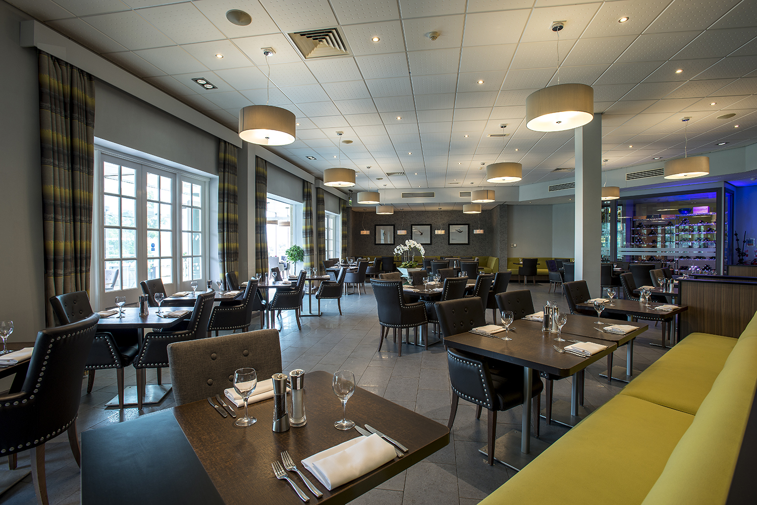 Modern restaurant interior with neatly arranged tables, upholstered chairs, and large windows. Ceiling lights hang above.