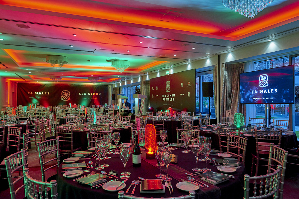 A dimly lit banquet hall with decorated tables, chairs, and screens displaying "FA WALES" and "CBD CYMRU" logos, in red and green lighting.