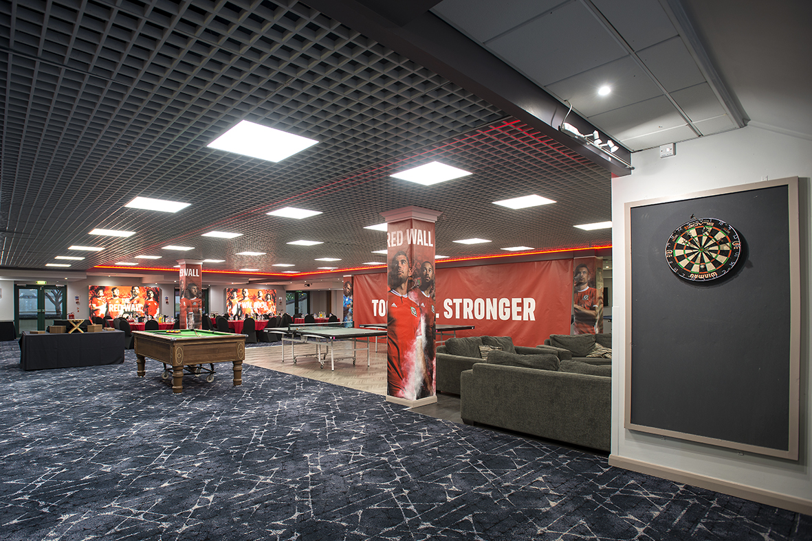 Lounge area with a pool table, dartboard, couches, and sports-themed decor featuring players and a "Red Wall" banner.