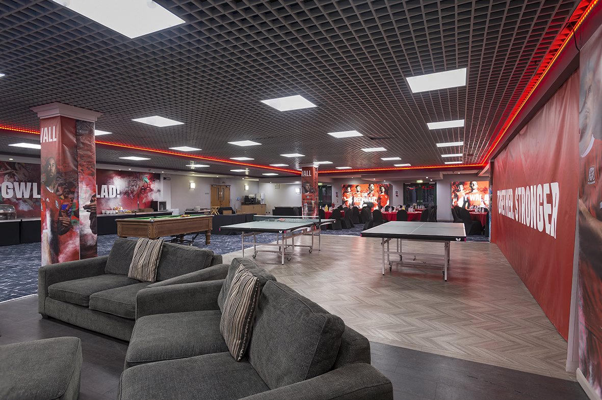 Spacious recreation room with sofas, pool table, ping pong tables, and red-lit decor, featuring sports-themed murals on walls.