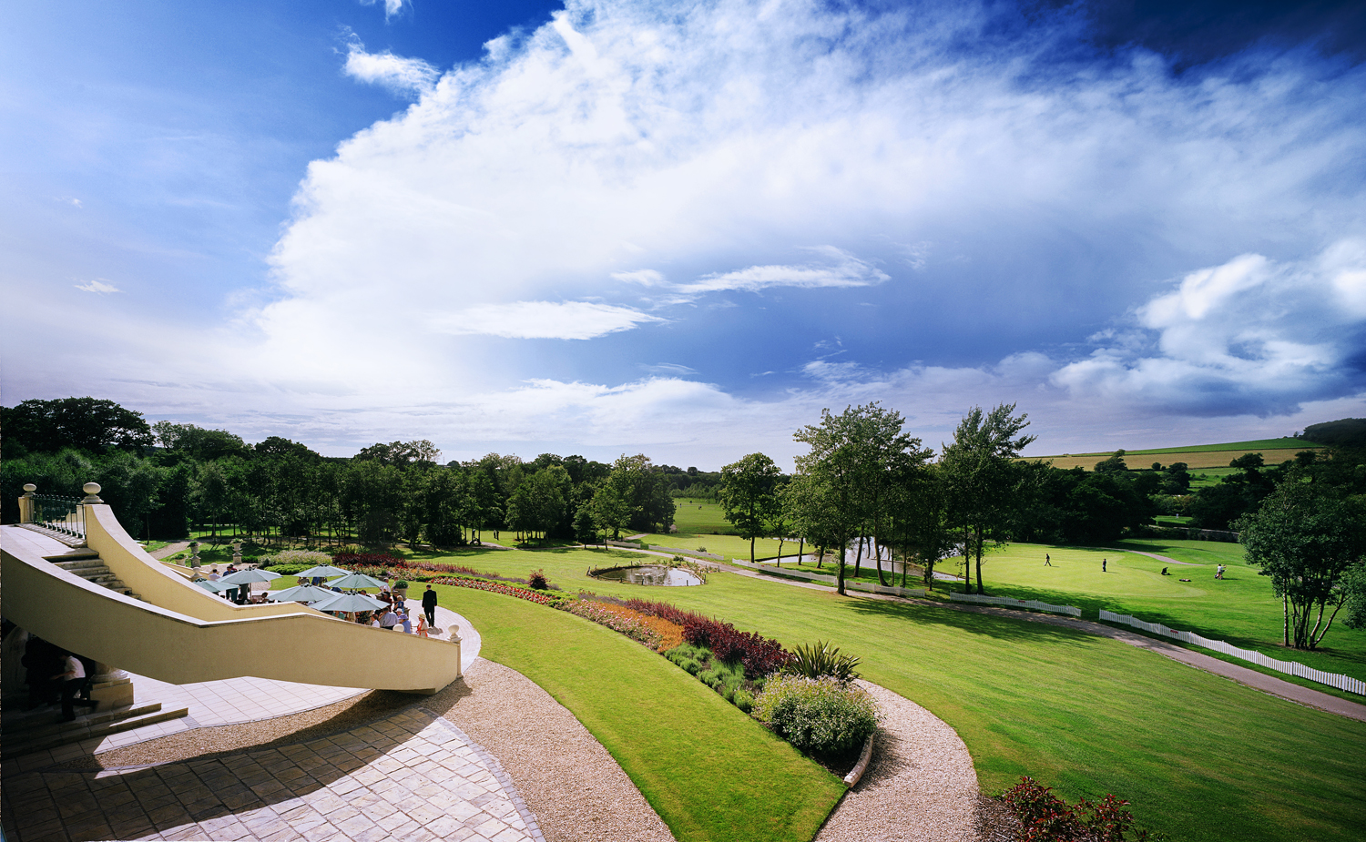A golf course with players on the green, surrounded by trees and rolling hills under a partly cloudy sky.