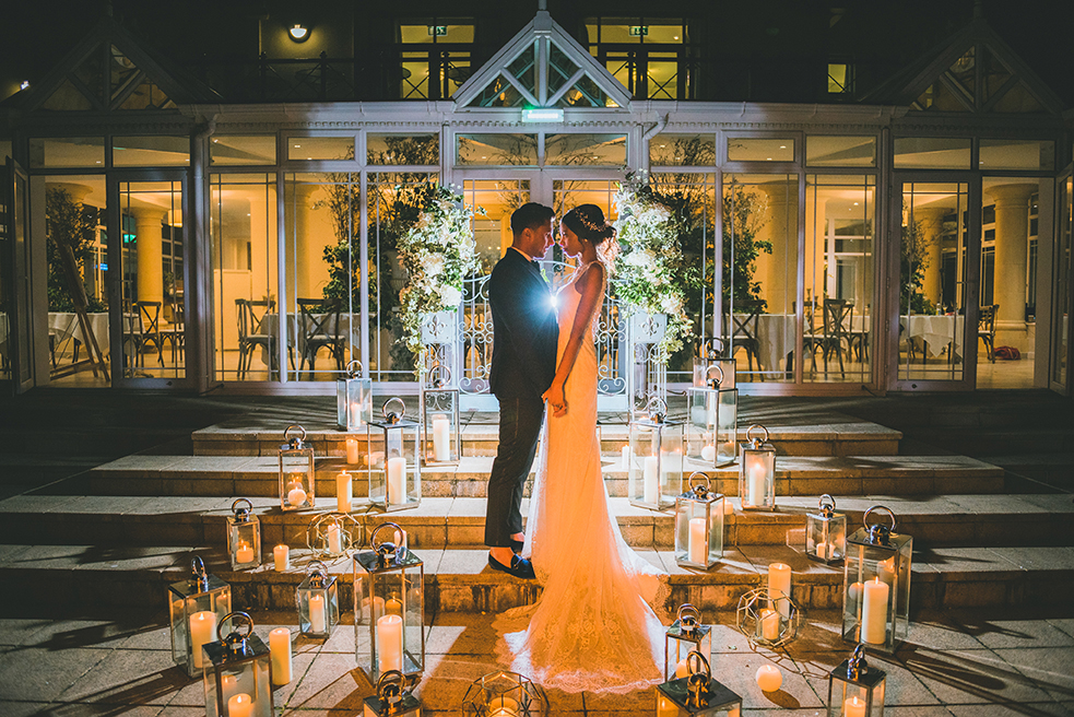 Bride and groom stand close together surrounded by lit candles and lanterns in front of a glass building at night.
