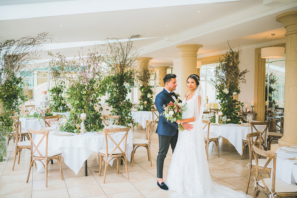 Bride and groom pose indoors at a wedding venue adorned with greenery and set tables, embracing each other warmly.