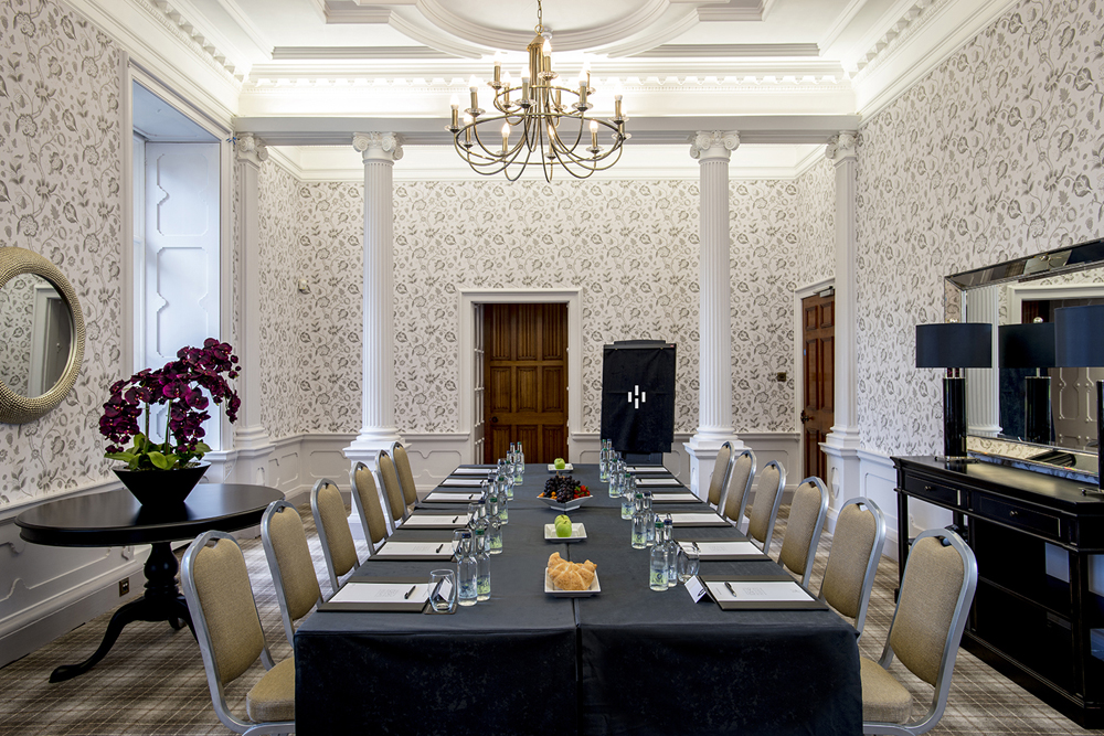 Elegant conference room with a long table set for a meeting, surrounded by chairs, floral centerpiece, and chandelier overhead.