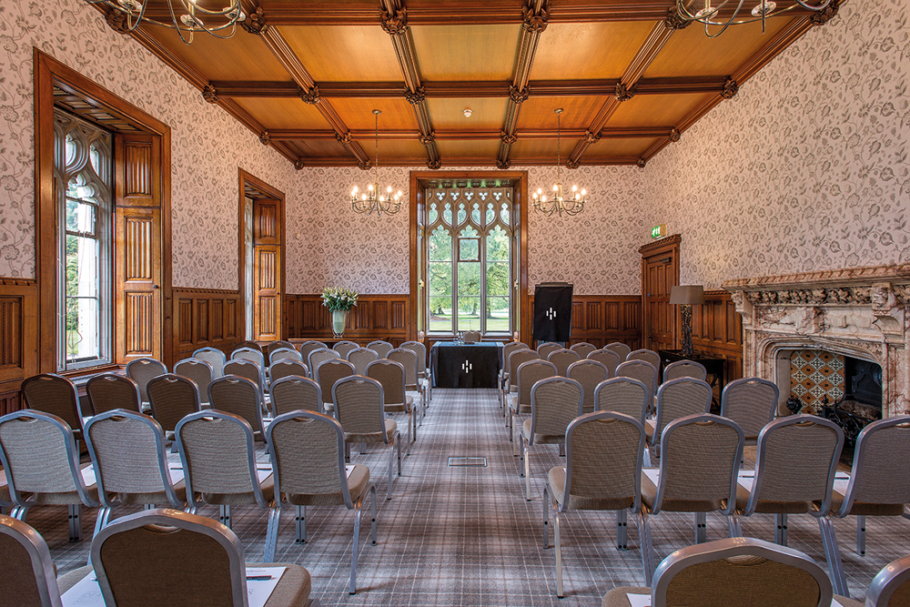 Room set up for a presentation with rows of empty chairs, wooden paneling, chandeliers, and a large window at the front.