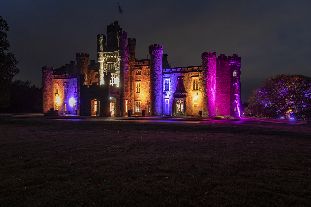 Illuminated castle at night with purple and orange lights, surrounded by dark trees and a lawn in the foreground.