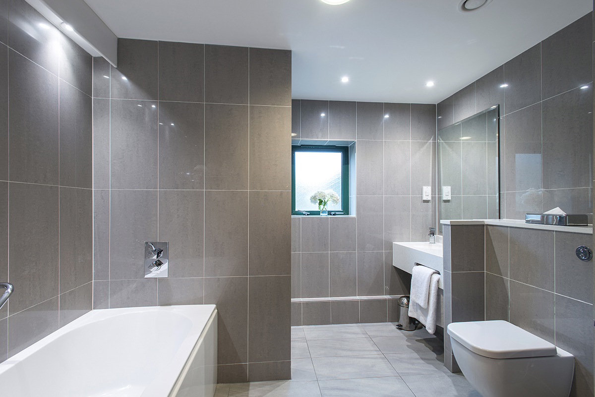 Modern bathroom with large gray tiles, a bathtub, and a wall-mounted toilet. A window and a sink with a mirror are visible.