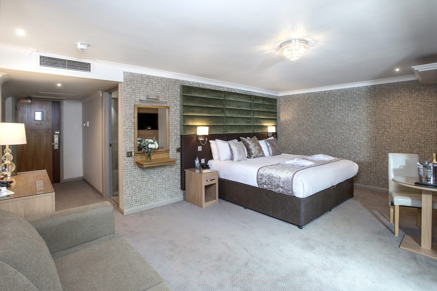 Spacious hotel room with a large bed, sofa, desk, and a small dining area. Neutral tones and soft lighting create a cozy feel.