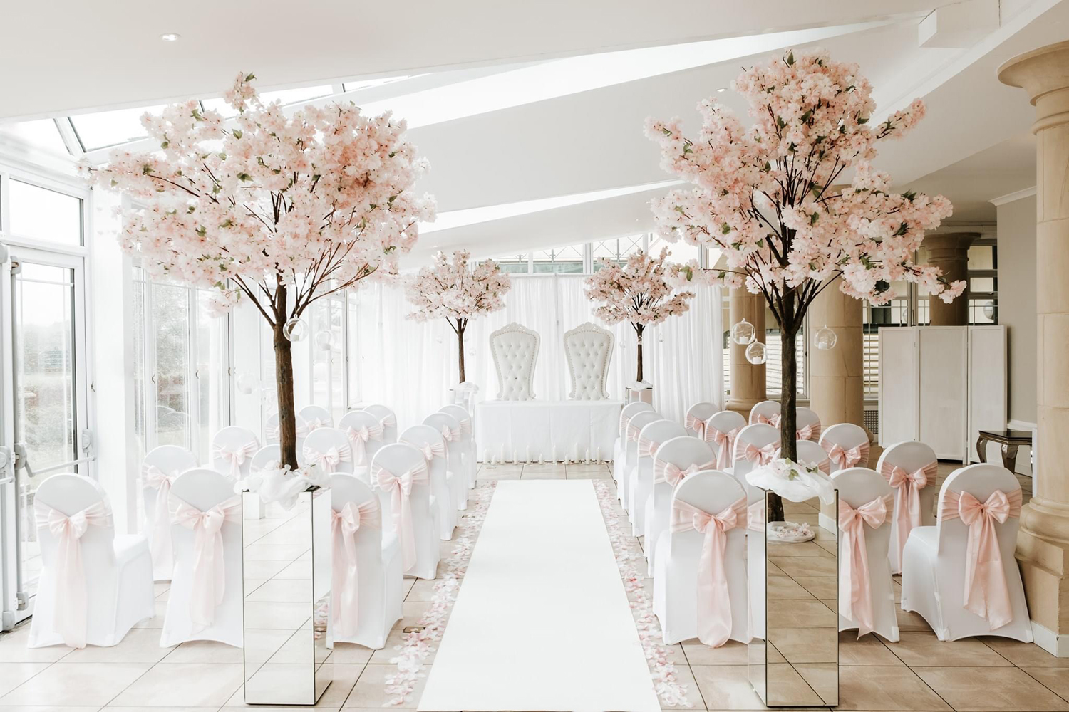 Elegant indoor wedding setup with white chairs decorated with pink bows, pink floral arrangements, and a white aisle runner.
