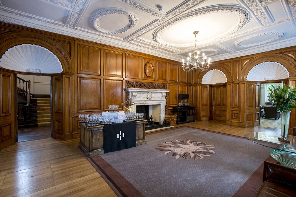 Elegant room with wood paneling, a decorative ceiling, a fireplace, and a table set with glassware on the floor.