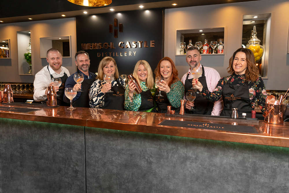 Seven people smiling and holding drinks at a bar in Hensol Castle Distillery, wearing aprons and standing behind a copper countertop.