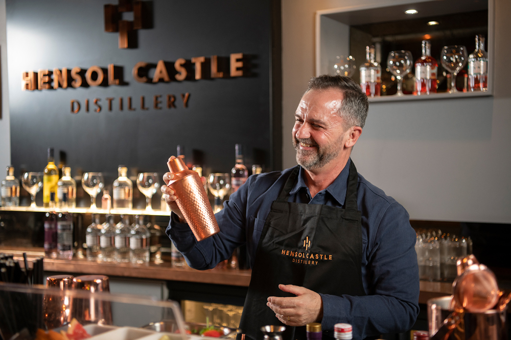 Man in a black apron holds a cocktail shaker at Hensol Castle Distillery, surrounded by bottles and glasses.