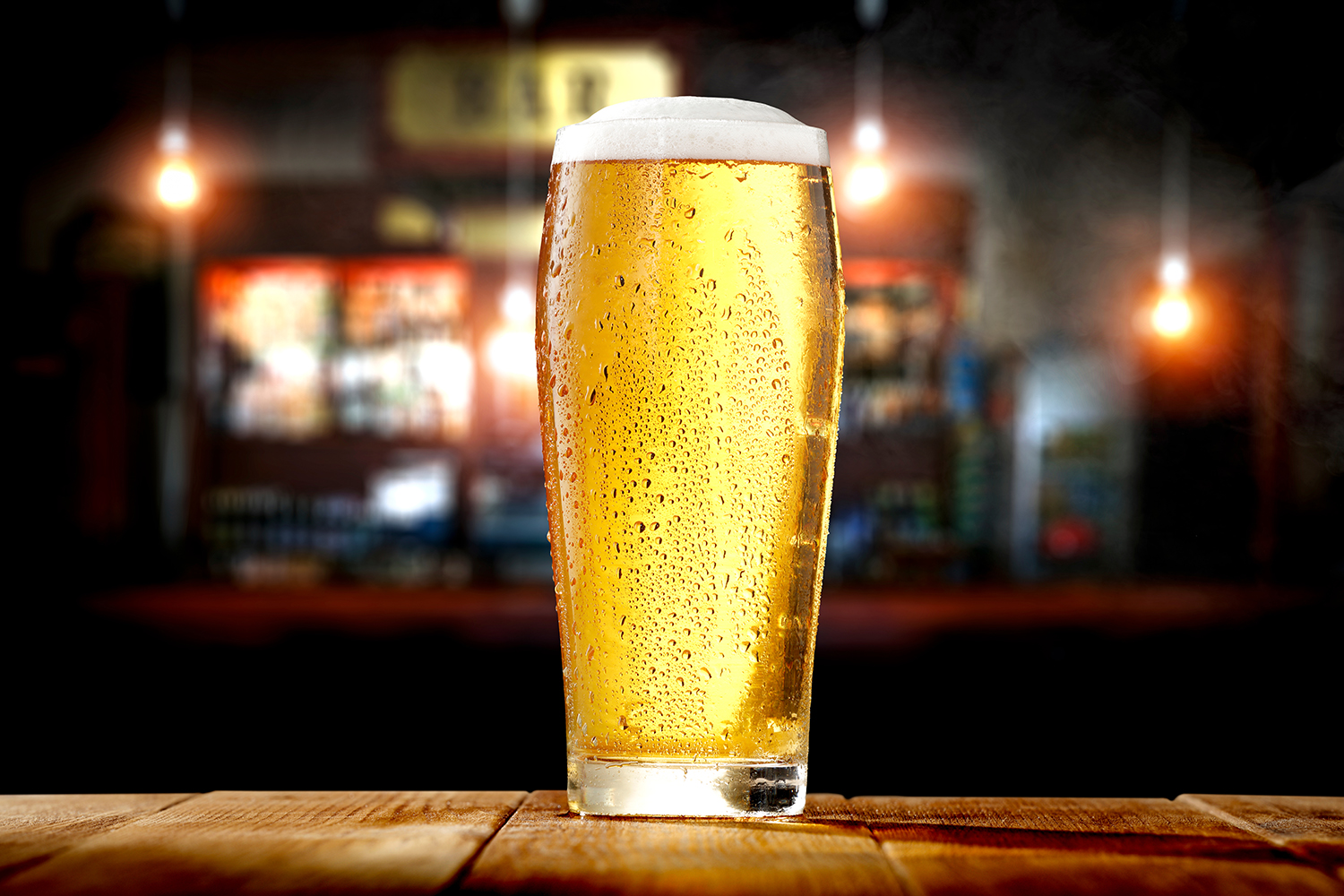 A pint glass of cold beer with condensation sits on a wooden surface, with a blurred bar background.