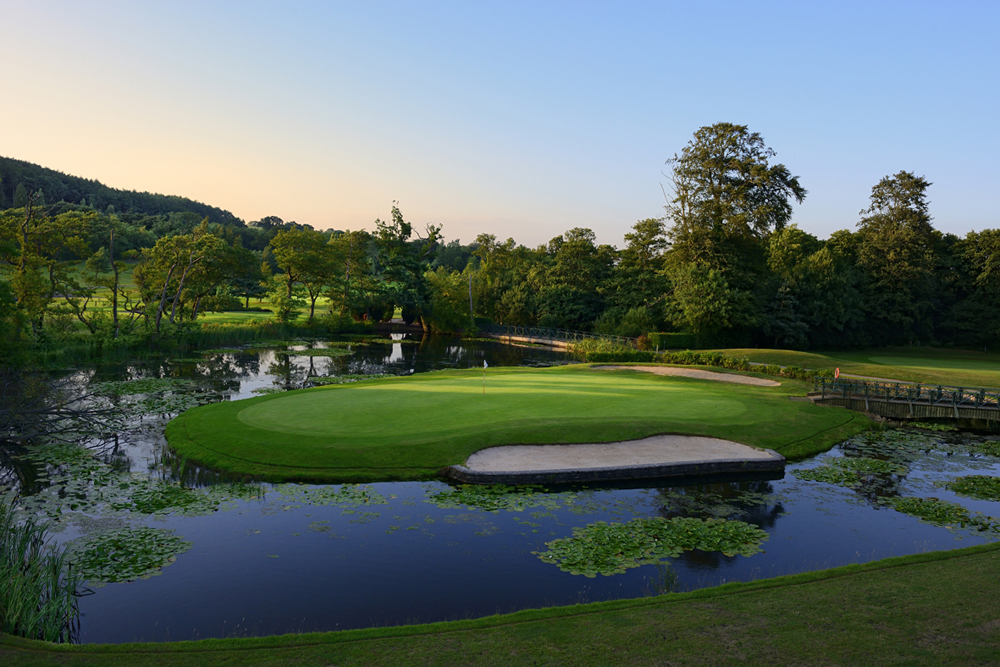 A serene golf course hole surrounded by water, with a sand bunker. Trees and blue sky are visible in the background.