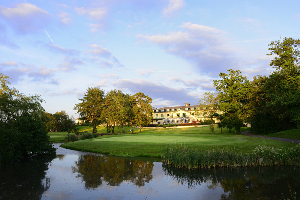 A large building sits on a lush green landscape with a pond in the foreground and a partly cloudy sky above.