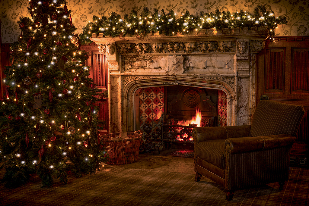 A decorated Christmas tree and a lit fireplace with a garland on the mantle in a warmly lit, cozy room.
