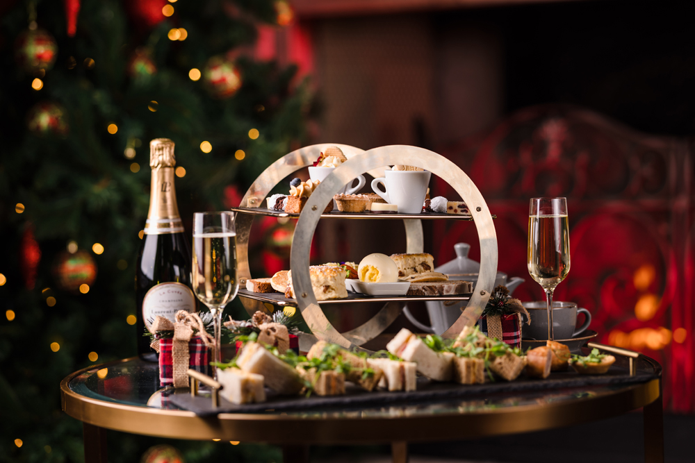 A table set with afternoon tea, sandwiches, pastries, champagne, and a Christmas tree in the background.