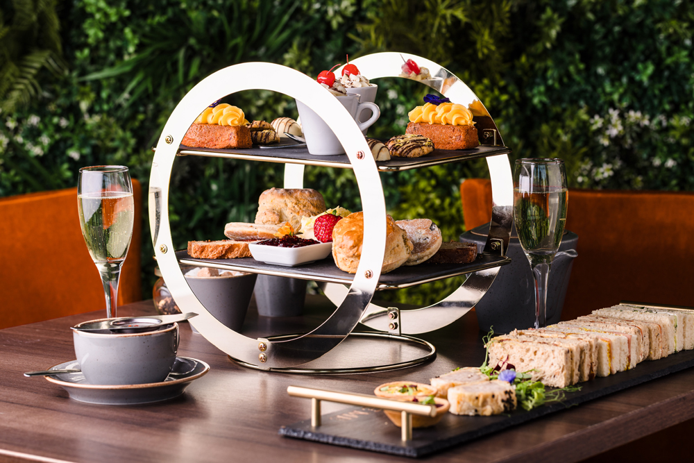 A modern two-tier afternoon tea stand with pastries, scones, sandwiches, and drinks on a wooden table.