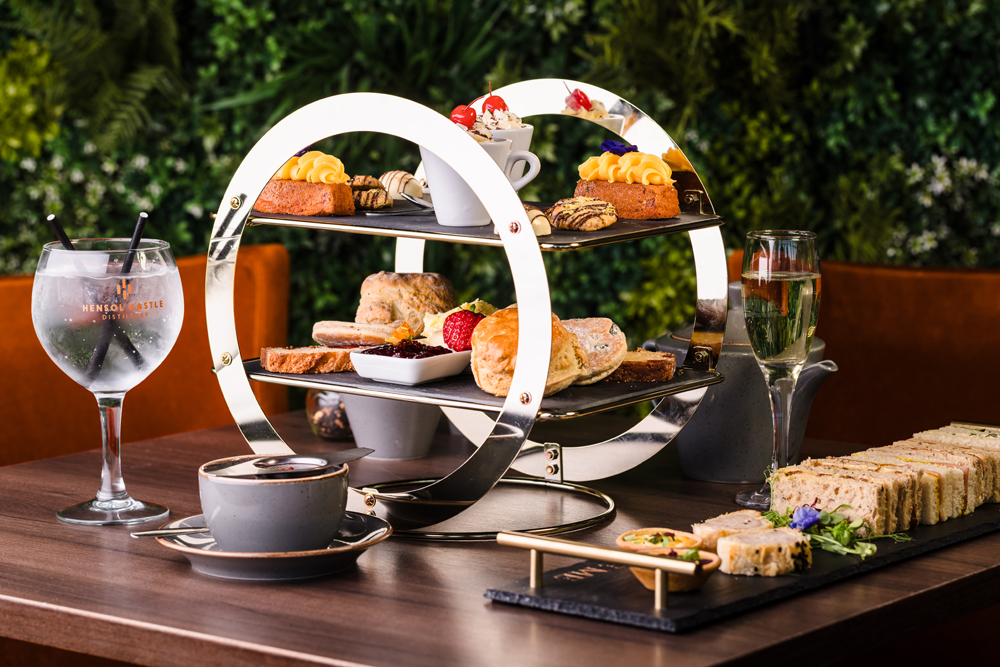 A tiered stand with pastries, scones, and desserts, alongside tea, sandwiches, a gin cocktail, and a glass of sparkling wine.