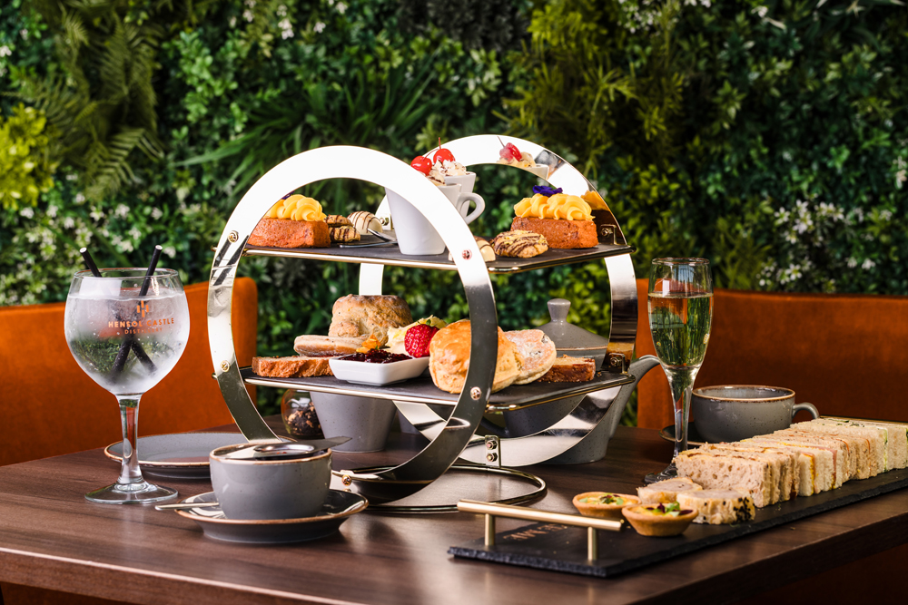 A table set with afternoon tea including pastries, scones, sandwiches, tea, a cocktail, and a glass of sparkling wine.