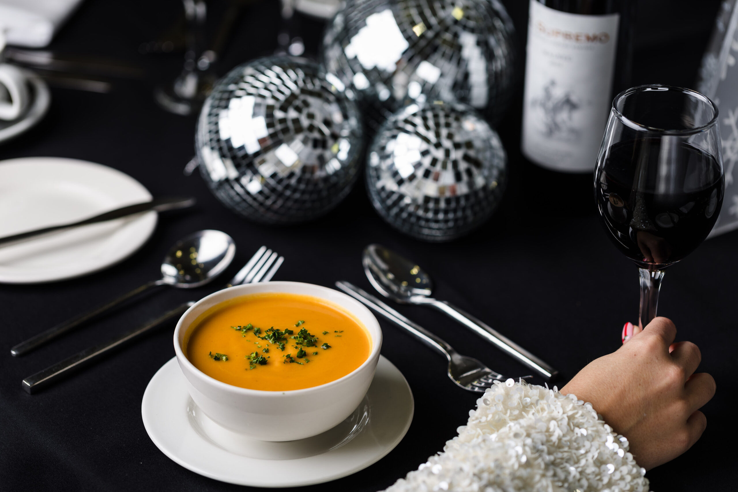 A bowl of soup on a table set with cutlery, disco balls, and a hand holding a glass of red wine.
