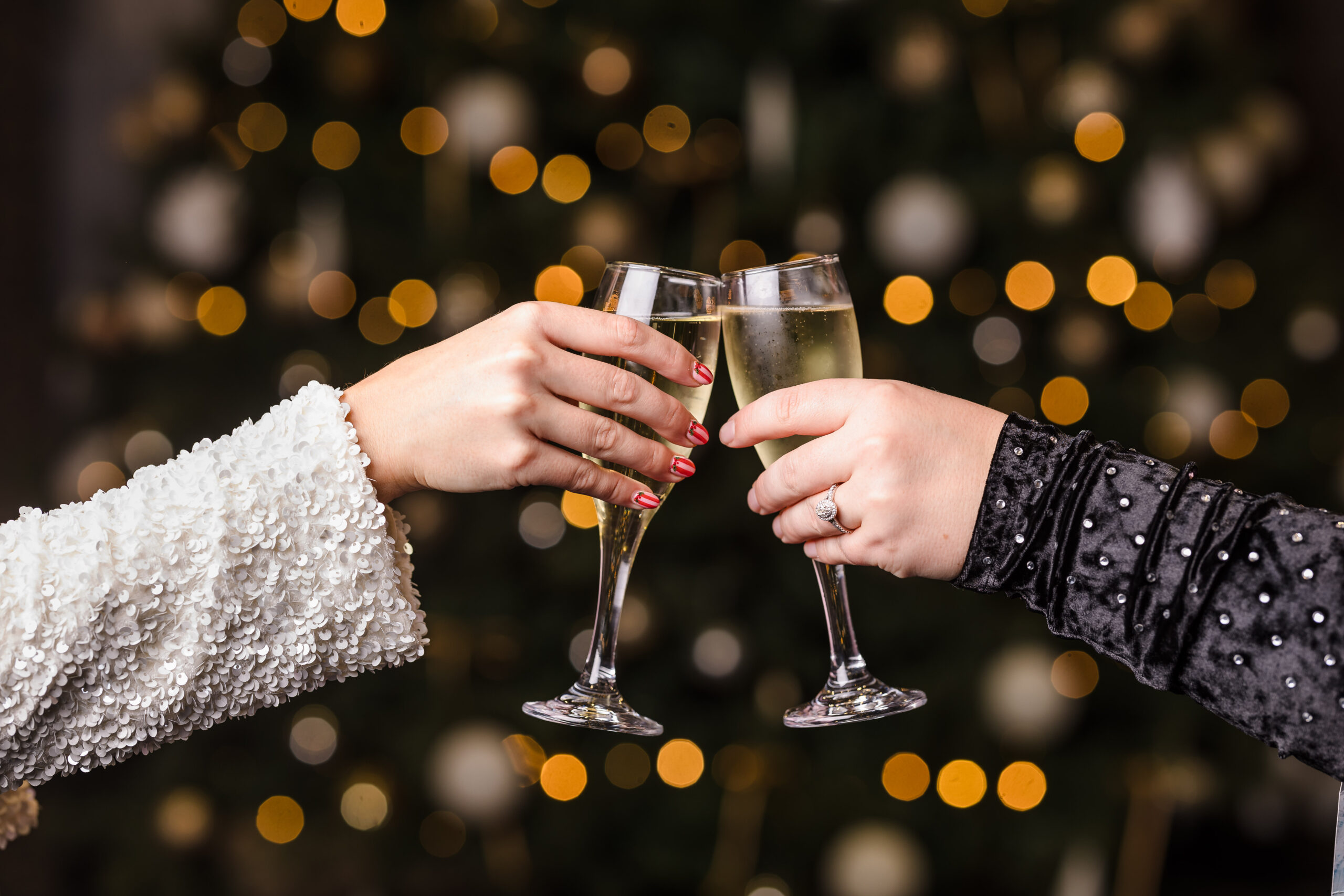 Two people clink champagne glasses in front of a decorated, blurred holiday tree with warm lights.