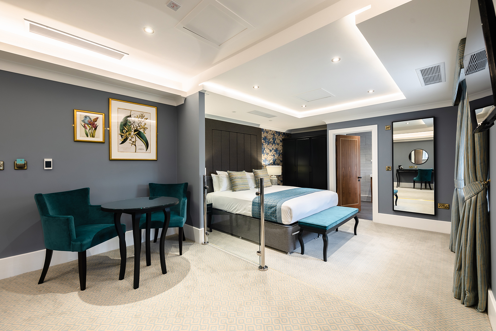 Modern hotel room with teal chairs, round table, large bed, wall art, and a floor-to-ceiling mirror.