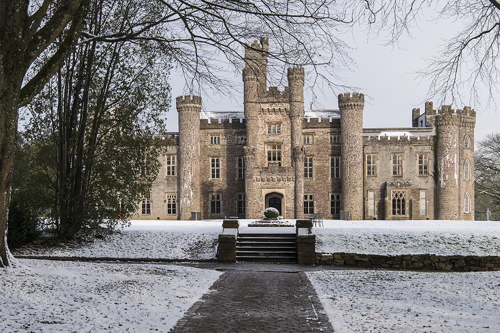 Stone castle with multiple towers and arched entrance, surrounded by trees and light snow on the ground.