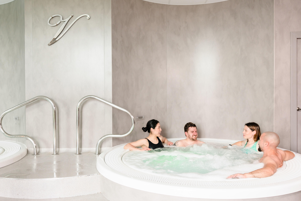 Four people sit and talk in a circular indoor hot tub next to metal stairs against a light-colored wall.