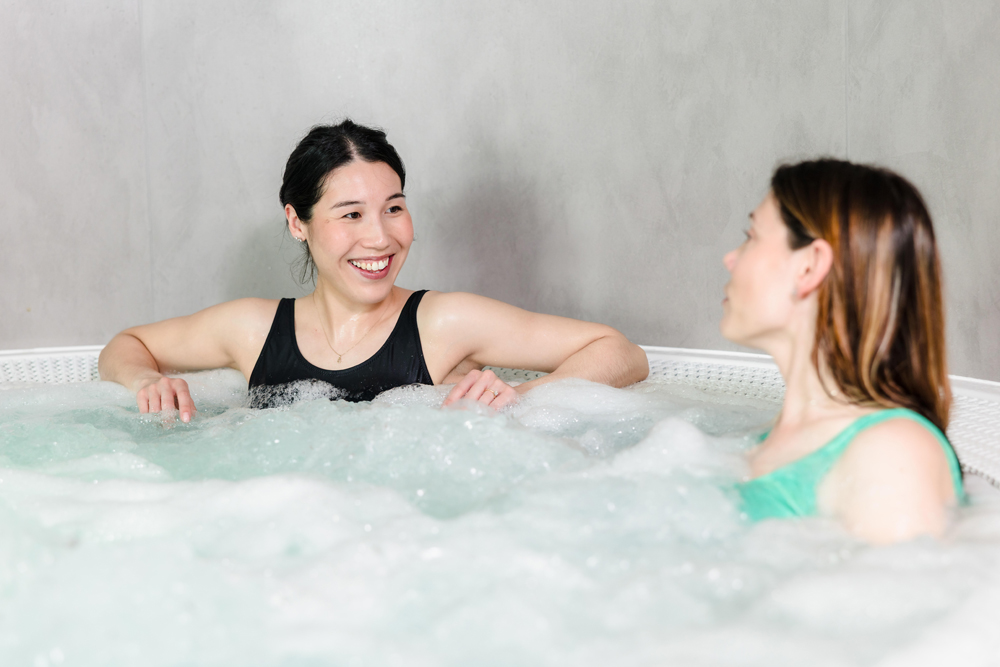 Two women wearing swimsuits sit and talk in a bubbling hot tub against a plain, light-colored wall.