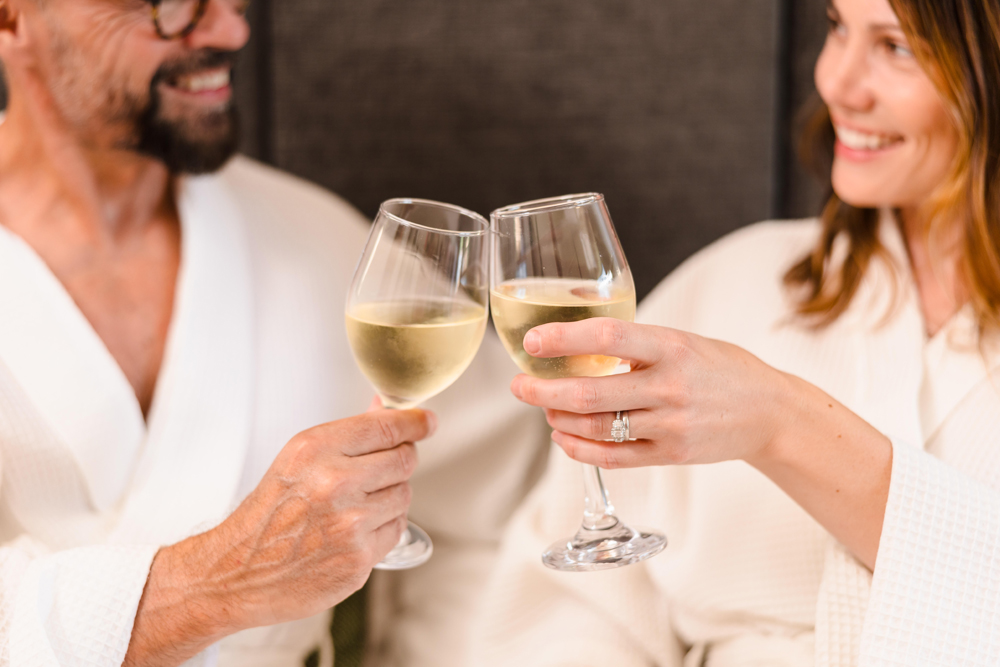 Two people in white robes clinking glasses of white wine and smiling at each other.