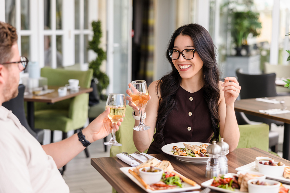 Two people sit at a restaurant table, smiling and clinking glasses of wine while enjoying a meal.