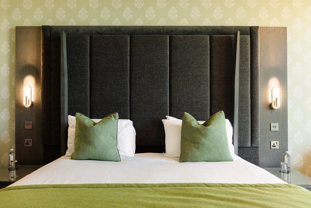 A neatly made bed with green pillows and a green throw, flanked by wall lights, against a patterned wallpaper.