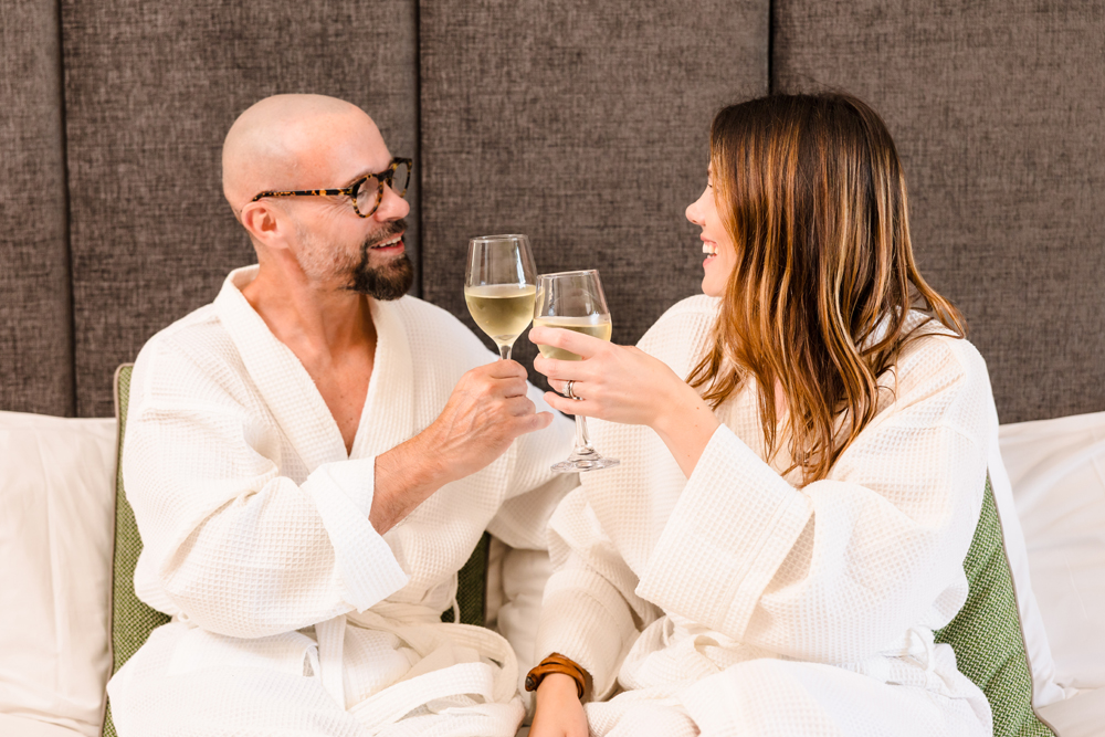 A man and a woman in white robes sit on a bed, clinking glasses of white wine and smiling at each other.