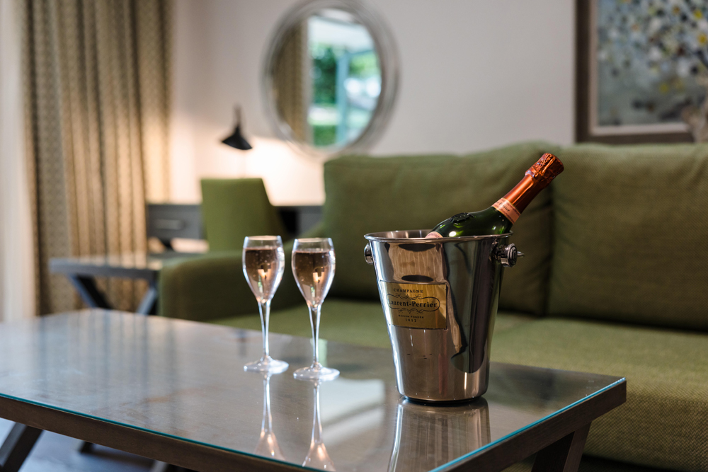 A bottle of champagne in an ice bucket and two filled glasses sit on a glass coffee table in a modern living room.