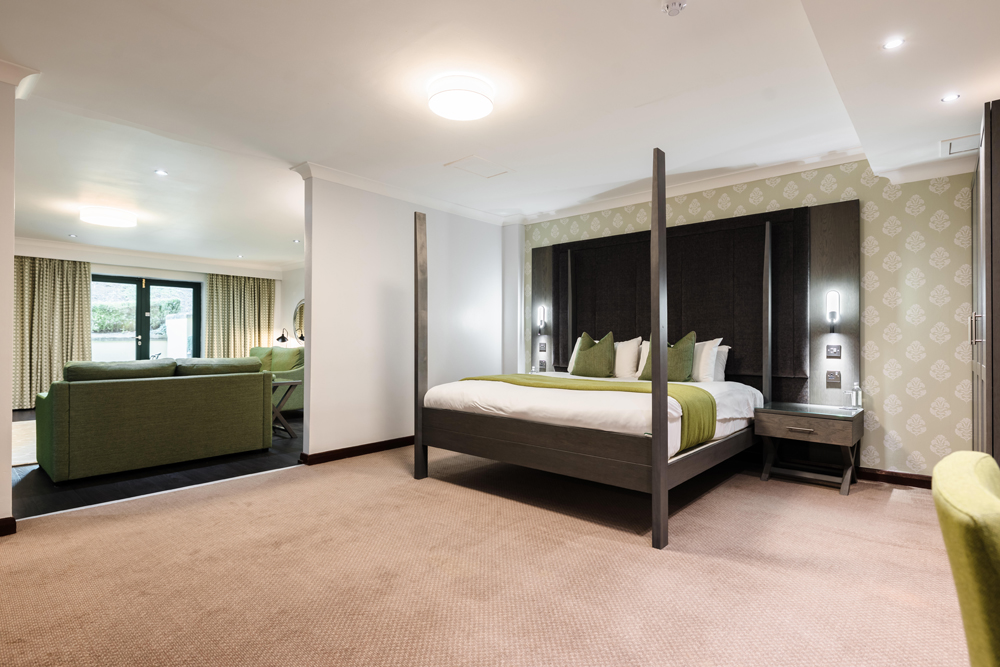 Spacious hotel room with a four-poster bed, nightstands, seating area, and large window with drapes.
