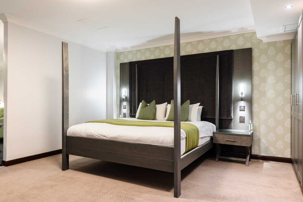 A modern bedroom with a four-poster bed, green accents, patterned wallpaper, and two bedside tables with lamps.