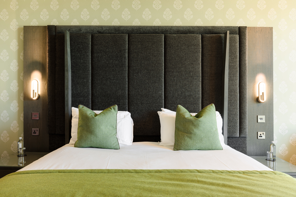 A neatly made hotel bed with green pillows and throw, a tall dark headboard, and two wall-mounted lights.